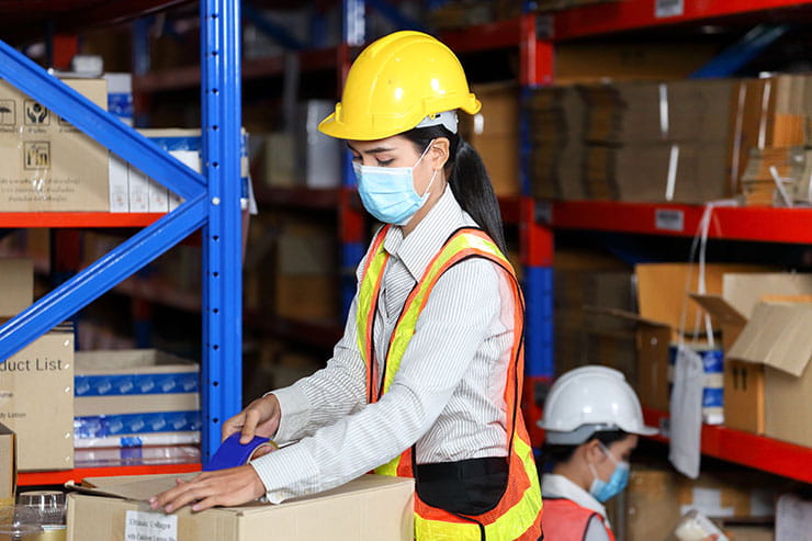Light industrial warehouse workers packaging boxes wearing surgical masks, helmets and safety vests while social distancing.